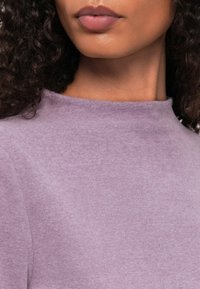 Paarse, gestructureerde stof met een hoge kraag. Dichtbijzicht van het kledingstuk, dat de zachtheid en subtiele kleurnuances benadrukt.