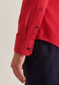 Chemise rouge à boutons avec un col pointu, dotée d'un bouton marine au poignet et d'un détail de rose brodé sur la manche.