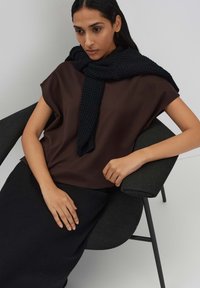 Braunes kurzärmliges Oberteil, das mit einem strukturierten schwarzen Schal geschichtet ist; Model sitzt in einem dunklen Stuhl und trägt einen langen schwarzen Rock.