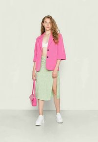 Blazer rosa brilhante sobre uma t-shirt cropped branca, combinado com uma saia estampada verde clara com fenda lateral. Os acessórios incluem uma mini bolsa rosa e ténis brancos.