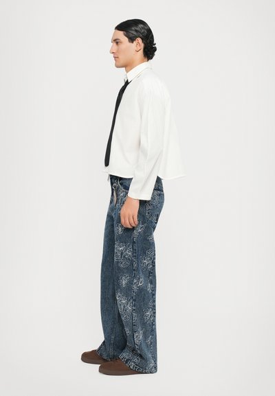 Camicia bianca a maniche lunghe con cravatta nera, abbinata a jeans in denim blu a gamba larga con ricami floreali bianchi e scarpe marroni scure.