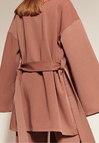 Woman wearing a loose, dusty rose jacket with wide sleeves and a self-tie belt at the waist, showing the back view.