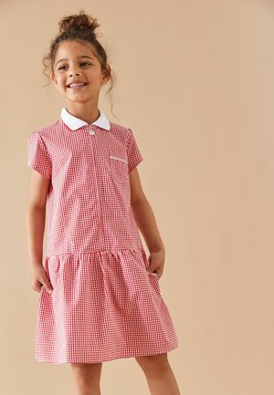 Lachend meisje met krullend haar in een knot, draagt een kort mouwtje rood-wit geruite jurk met een witte kraag tegen een beige achtergrond.