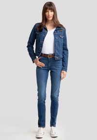 Traper jakna s plavom tkaninom i frontom na dugmad, uparena sa slim fit plavim trapericama i bijelom majicom. Bijele tenisice upotpunjuju odjevnu kombinaciju.