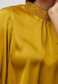 Blouse jaune moutarde avec un col froncé et une texture douce et lisse. Dotée de manches fluides et de plis pour une coupe décontractée.
