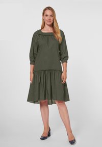 Dunkelgrünes Kleid mit quadratischem Ausschnitt, dreiviertel langen Ärmeln und einem gerafften Rock. Hergestellt aus leichtem Stoff mit lockerer Passform.