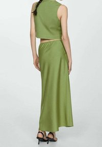 Frau mit geflochtenem Haar, die ein ärmelloses grünes Crop-Top und einen passenden langen Rock trägt, schwarze High Heels, steht mit dem Rücken zur Kamera.