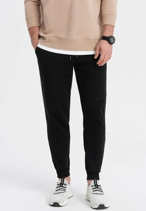 Sorte joggerbukser med en tapered design, der har en elastisk talje og sidelommer. Parret med hvide træningssko og en beige top.