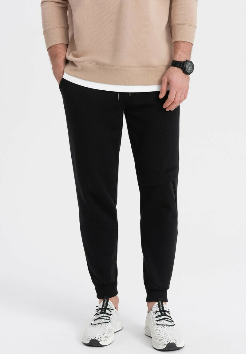 Schwarze Jogginghose mit tailliertem Design, ausgestattet mit einem elastischen Bund und einer Seitentasche. Kombiniert mit weißen Sportschuhen und einem beigen Oberteil.