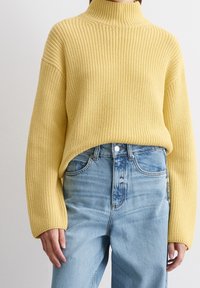 Żółty sweter z prążkowanej dzianiny z wysokim kołnierzem i luźnymi długimi rękawami, połączony z jasnoniebieskimi dżinsami o wysokim stanie.