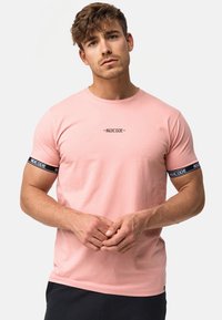 Tricou din bumbac roz deschis, cu decolteu rotund, mâneci scurte și accente de bandă neagră cu text. Textură netedă.