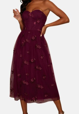 Vestido midi burdeos sin tirantes con bordado floral, corpiño ajustado con varillas y falda superpuesta transparente, llevado por una mujer con una mano en la cadera.