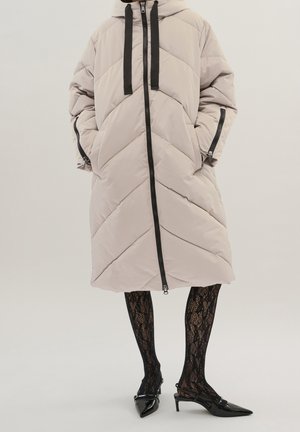 Personne portant un long manteau matelassé beige avec fermeture éclair et poignets noirs, associé à des collants noirs à motif dentelle et des talons pointus noirs.