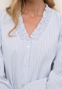 Blouse à rayures bleues et blanches avec un col en V volanté et des manches longues. Tissu doux avec des rayures verticales et fermeture par bouton.