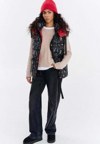 Czarna pikowana kamizelka z kwiatowym wzorem, czerwonymi akcentami i wysokim kołnierzem, noszona na beżowym, dzianym swetrze; zestawiona z luźnymi czarnymi dżinsami i czarnymi botkami.