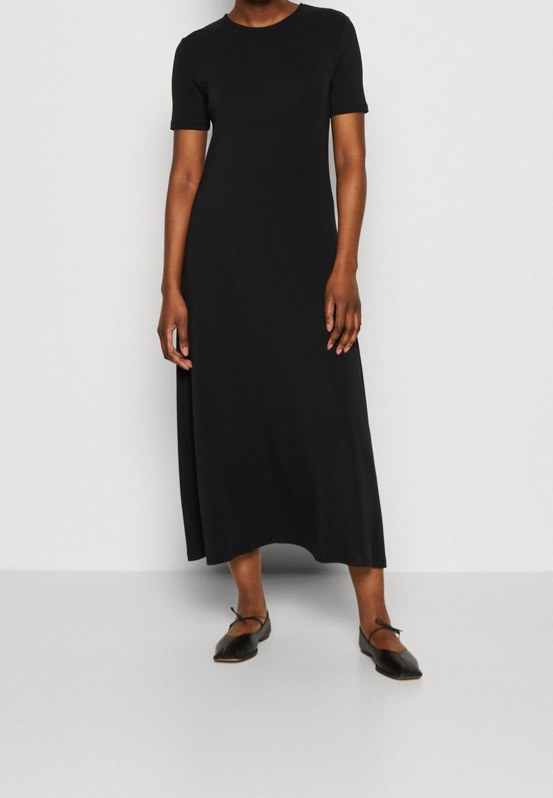 Femme portant une robe midi noire unie à manches courtes et des ballerines noires avec des brides à la cheville, debout contre un mur blanc.