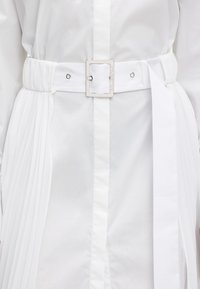 Fehér ing, amihez összegyűrt fehér szövet öv tartozik. Négyzet alakú ezüst csattal és textil díszítéssel rendelkezik az övön. Az egész sima tapintású.