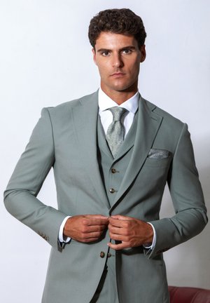 Mann, der einen hellgrauen Dreiteiler mit Paisley-Krawatte und passendem Taschentuch zuknöpft, steht vor einem schlichten weißen Hintergrund.