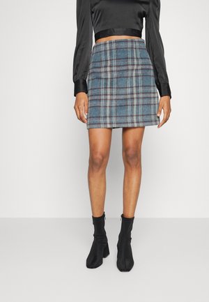 Frau trägt einen hoch taillierten blauen Minirock mit Karomuster, eine schwarze Satinbluse mit langen Ärmeln und schwarze Knöchelhohe Stiefel, steht vor einem weißen Hintergrund.