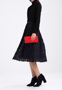 Zwarte top met lange mouwen, marineblauwe kanten rok, gestructureerde rode clutch en zwarte enkellaarsjes. De clutch heeft een geometrisch ontwerp.