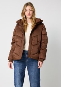 Giubbotto imbottito marrone con cappuccio, caratterizzato da un design trapuntato, bottoni a scatto e polsini a costine. Indossato sopra una maglietta beige chiaro e un paio di jeans blu.