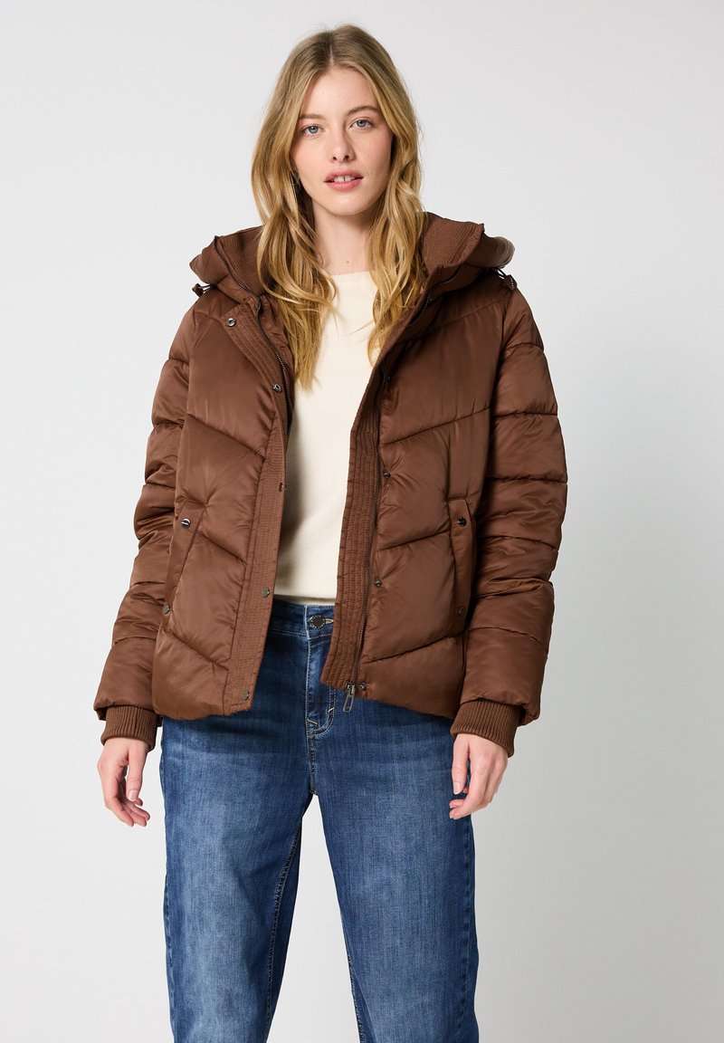 Giubbotto imbottito marrone con cappuccio, caratterizzato da un design trapuntato, bottoni a scatto e polsini a costine. Indossato sopra una maglietta beige chiaro e un paio di jeans blu.