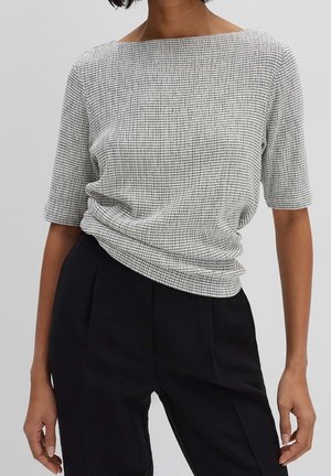 Femme portant un haut blanc à motif quadrillé avec des manches aux coudes et un pantalon noir taille haute sur un fond uni.