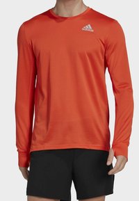 T-shirt de sport à manches longues de couleur orange vif, fabriqué en tissu texturé. Il est doté d'un col rond et d'un petit logo Adidas blanc sur la poitrine.
