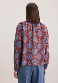 Blusa a maniche lunghe in tessuto rosso con motivi floreali blu. Presenta un colletto arricciato e polsini con elastico. Abbinata a un jeans loose.