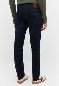 Ciemnoniebieskie dżinsy o dopasowanym kroju, wykonane z denimu; posiadają pięć kieszeni, subtelne przeszycia oraz brązową skórzaną naszywkę z logo na pasku.