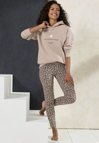 Sudadera beige con el texto "HACES SONREÍR A MI CORAZÓN" y un corazón dorado. Leggings ajustados de estampado de leopardo en beige y negro. Modelo posando en unas escaleras.