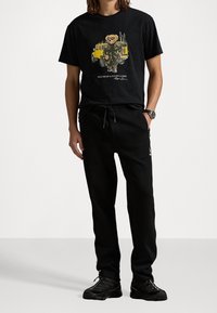 Czarna bawełniana koszulka z kolorową grafiką niedźwiedzia, w połączeniu z czarnymi spodniami dresowymi. Stylizacja dopełniona czarnymi sneakerami.