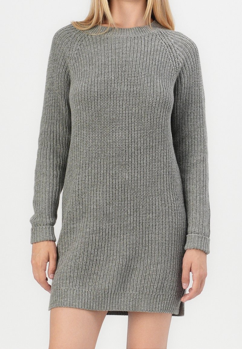 Femme portant une robe-pull en maille grise à manches longues avec des poignets et un ourlet côtelés sur un fond blanc uni.