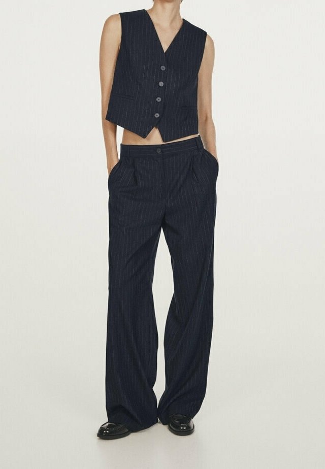 Personne portant un gilet sans manches noir à fines rayures boutonné et un pantalon large assorti, mains dans les poches et chaussures noires.