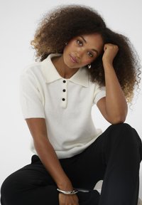 Polo shirt din tricot alb, cu mânecă scurtă, căptușit cu nasturi negri și un guler în contrast. Țesătura pare moale, cu o textură fină. Pantaloni negri sunt vizibili.