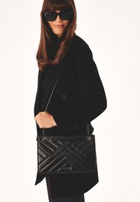 Borsa a tracolla in pelle nera con cuciture a zig-zag, tracolla in catena e dettaglio del logo. Indossata con un cappotto nero, che mette in evidenza un design elegante e strutturato.