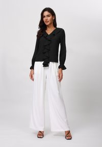 Schwarze Bluse mit Rüschendetail vorne und langen Ärmeln, kombiniert mit weißen, weit geschnittenen Hosen, getragen mit schwarzen Absatzsandalen.