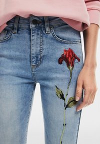 Vaaleansiniset farkut kapealla räätälöinnillä, joissa on punainen ruusu brodeerattu vasempaan lahkeeseen, ja vihreitä lehtiä sekä mustia yksityiskohtia.