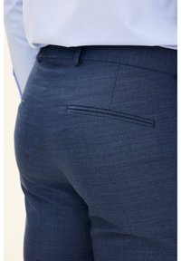 Marineblaue Hose aus strukturiertem Stoff. Verfügt über eine Gesäßtasche mit einer einzelnen horizontalen Naht. Kombiniert mit einem hellblauen Hemd.