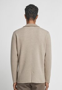 Beige Strickpullover mit Schalkragen, langen Ärmeln und einer mittigen Rückennaht. Weicher Stoff, lässige Passform und seitlicher Schlitz am Saum.