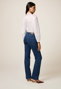 Bluză albă ușoară cu detalii din dantelă pe spate, asortată cu blugi drepti de culoare albastră închis, având o etichetă din piele maro.