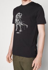 Svart t-shirt med ett vitt mönster av en kretskort som formar en T-Rex och ordet "BOSS" tryckt nedanför på en stående person.