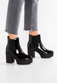 Černé boty kotníkové z lakované kůže s robustními podpatky a gumovými podrážkami, vybavené elastickými bočními panely a texturovanými detaily.