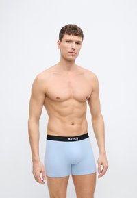 Ευθυτενής άνδρας που στέκεται μπροστά από απλό φόντο, φορώντας ανοιχτό μπλε ανδρικά εσώρουχα BOSS με μαύρη μέση, κοιτώντας μπροστά με ουδέτερη έκφραση.