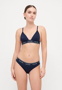Námornícky trojuholníkový bralet a zodpovedajúce bikiny. Mäkka tkanina s pásom s logom Calvin Klein. Nastaviteľné ramienka, minimalistický dizajn.