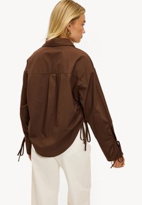 Chemise marron légère à coupe ample, avec manches longues, détails noués sur les côtés et ourlet arrondi.