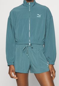 Letvægts træningssæt i grønblå med lynlåsjakke og elastik i taljen. Jakken har en høj krave, mens shortsene har sidelommer og snor i taljen.