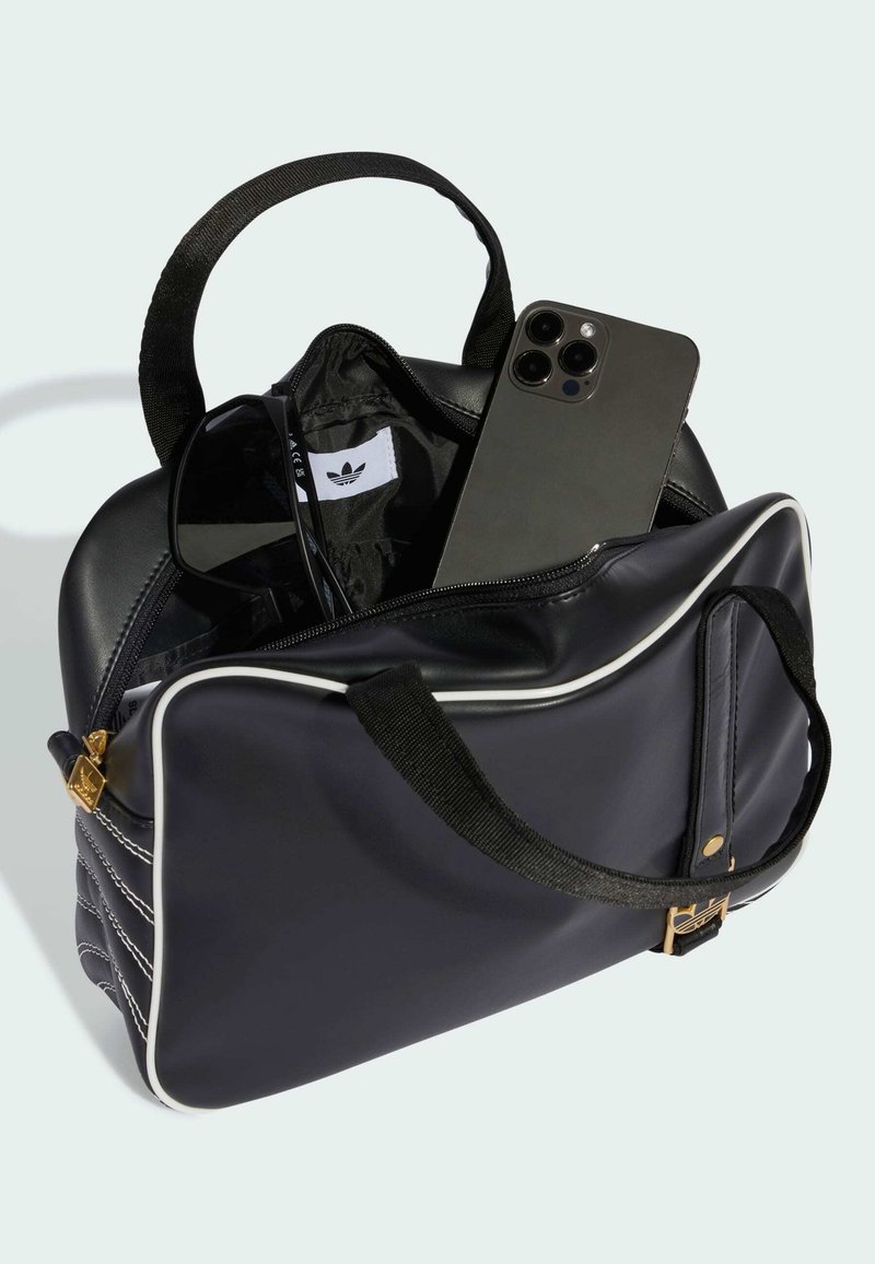 Sac de sport noir Adidas avec fermeture éclair dorée, lunettes de soleil noires et un smartphone partiellement visible à l'intérieur sur fond clair.