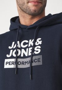 Námornícky modrá mikina s kapucňou a šnúrkami, s výrazným bielym textom: "JACK & JONES PERFORMANCE." Mäkko tkanina a štandardný strih.