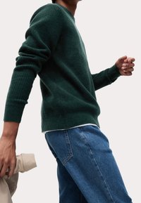 Ciemnozielony, teksturowany sweter z długimi rękawami, noszony na białej koszuli. W połączeniu z niebieskimi dżinsami, postać jest widoczna z profilu.